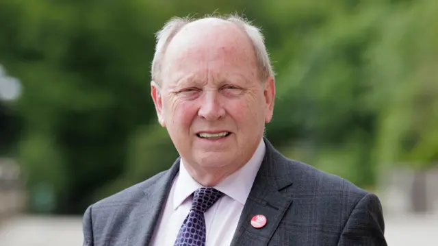 A man stares at the camera. He is wearing a grey suit with a white shirt and a purple tie. Behind him, out of focus, there are trees.