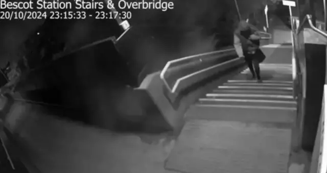 A black and white CCTV image of a man with a hood up walking up steps at a bridge over a railway line