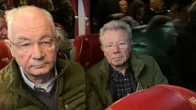 Two men sitting together on a coach. They both have short grey hair. They have green jackets on.