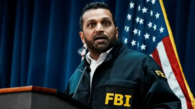 FBI Director Kash Patel stands in front of an American flag while wearing a navy blue jacket with large yellow letters on the front pocket and sleeve saying 'FBI'