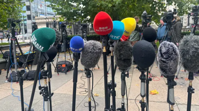 A group of microphones on stand, there are camera people and cameras in the background