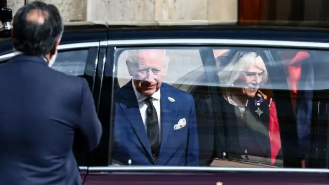 King Charles and Camilla enter black car, they are seen through the window. Charles wears a blue suit as Camilla is in a black jacket.