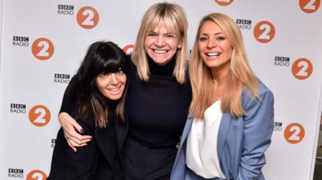Claudia Winkleman, Zoe Ball, and Tess Daly post in front of a Radio 2 poster