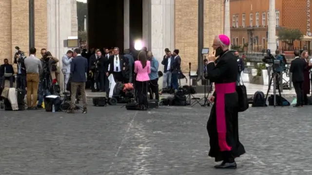 A group of press with cameras and lights stand outside building in the Vatican - a Cardinal walks past on the phone.