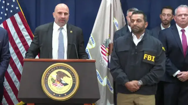 Christopher Raia speaking at podium that says Department of Justice, next to him is Kash Patel - with FBI written on his jacket in large yellow letters.