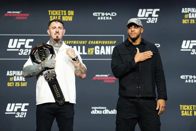 Tom Aspinall holds up his fist and has the UFC heavyweight title on his shoulder and stands next to Ciryl Gane who is placing his hand on his chest