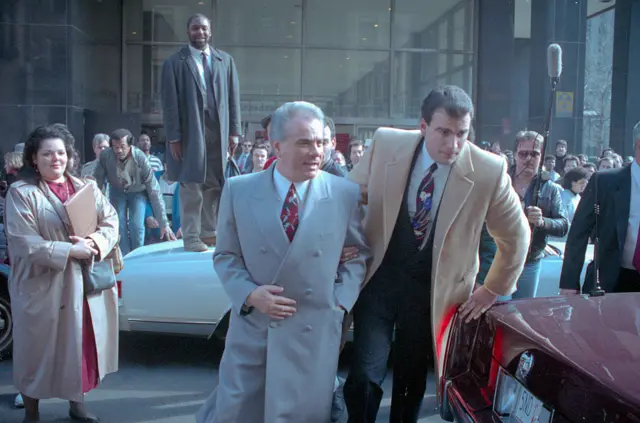 Gotti walking in very dapper suit and tie as press crowds behind him