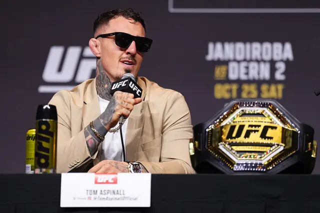Tom Aspinall speaks into a microphone and sits next to the UFC heavyweight title