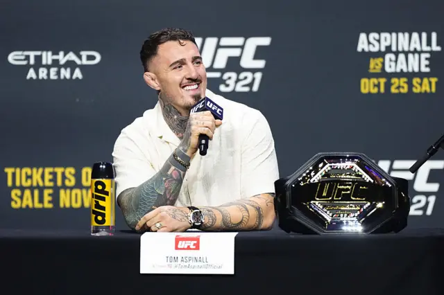 Tom Aspinall holds a microphone to his mouth and has the UFC heavyweight title next to him on a table
