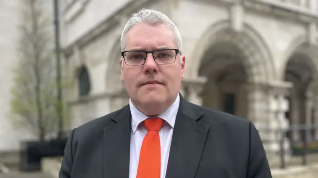 A man with short white hair and glasses looks at the camera. He is wearing a dark suit and a bright red tie.
