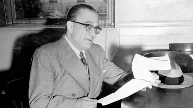 Genovese wearing a suit sits at a desk looking through papers