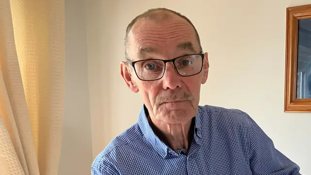 A man, with balding grey hair, wearing a blue and white check shirt and black rimmed glasses.