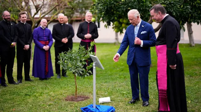 King Charles in a blue suit stands next to a man wearing black robes, they look at a plaque and a tree