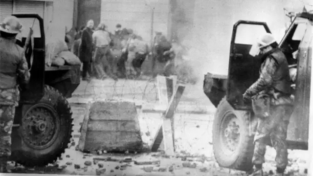 A black and white photograph taken on Bloody Sunday. There are two soldiers in the foreground, standing behind their sandbagged armoured car. There is a barbed wire barricade in front of them and in the further distance, there is a group of people with their backs towards them in the opposite direction. There is rubble on the ground and smoke in the air.