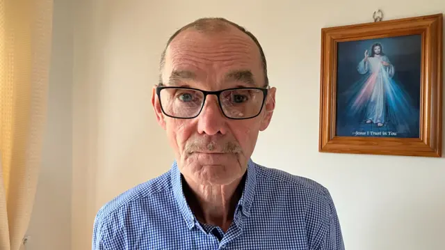 An old man wearing glasses looks directly at the camera. He is wearing a blue and white shirt. Behind him is a framed portrait of Jesus.