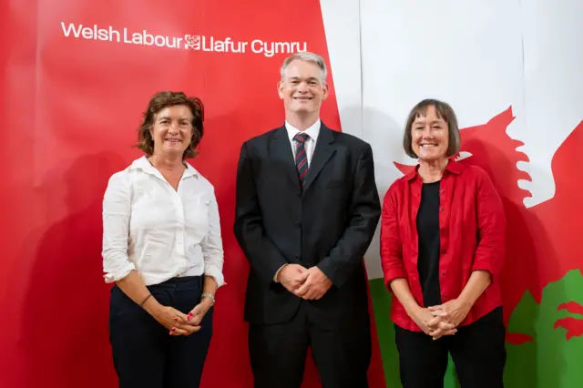 Eluned Morgan, Richard Tunnicliffe and Jo Stevens stood in front of a Welsh flag and a Welsh Labour sign