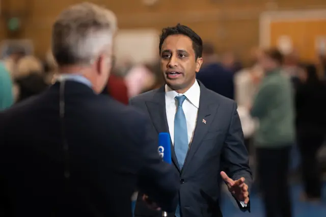 Zia Yusuf talking to a reporter. He has a blue suit, white shirt and blue tie.