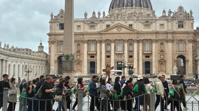 A group of people walk in front of St Peter's Basilica in Vatican City, a large barrier in the foreground.