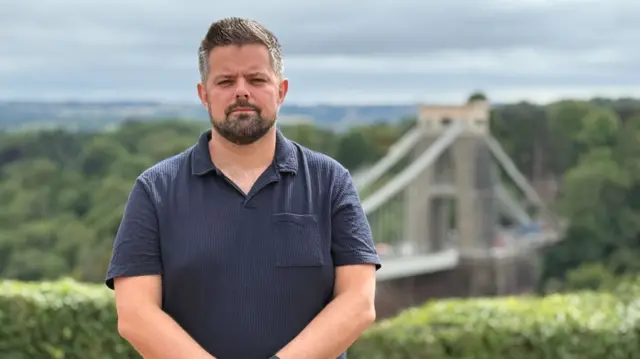 Bearded male presenter stands with the Clifton Suspension Bridge in the background.
