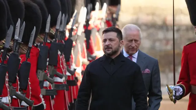 Zelensky dressed in black inspects the troops in red uniform and black bearskin hats as King Charles walks behind him