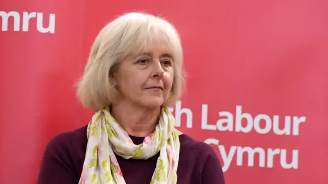 Ruth Jones looking to the right, she has shoulder-length grey hair and is wearing a plum top and a white, yellow and pink scarf. Behind her is the Welsh Labour red board