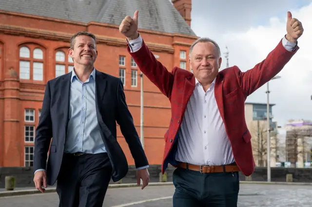 Photograph of Rhun ap Iorwerth (left) and Lindsay Whittle (right) walking into the Senedd building in Cardiff bay. Iorwerth wears a two piece navy suit and blue shirt and smiles widely. While Lindsay wears a deep red check blazer with a white shirt. He holds both his arms above his head in fights and smiles.