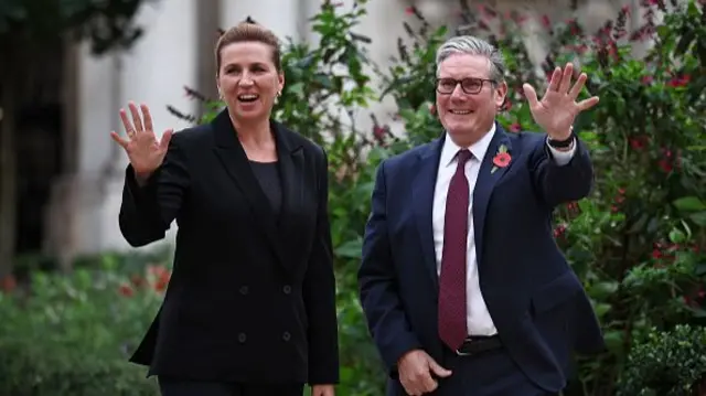 A woman in a black blazer waves next to a man in a blue suit