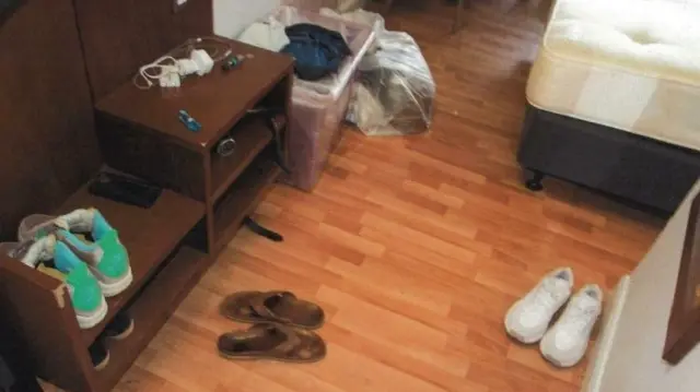 A police image inside a hotel room with wooden floors. There is a pair of brown flip flops on the ground and a pair of brown trainers. in the background you can see a bed and bags of rubbish in clear bags