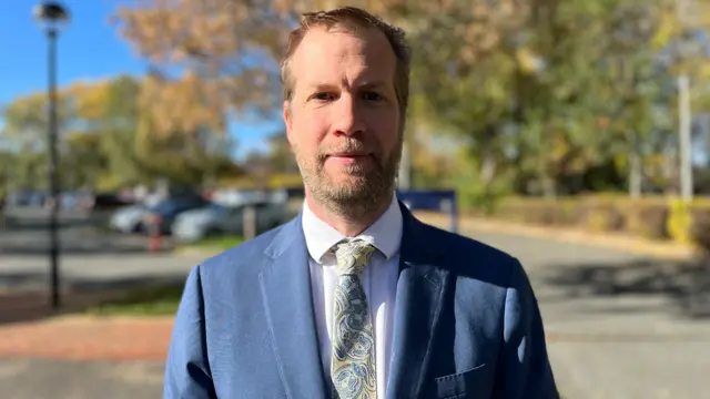 Det Ch Insp Stride looks at the camera with a car park and trees behind him. He has a short beard and short blonde hair, and wears a blue suit with a yellow and blue paisley tie.