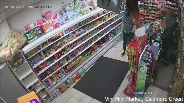 CCTV image inside a shop with aisles stacked with refrigerated drinks. A man in a grey and silver top is walking in, with white earbuds in his ears