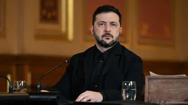 Zelensky in black blazer and black shirt sits down at table during coalition of the willing meeting at the FCDO