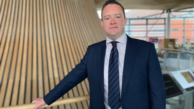 Gareth Davies, Welsh conservative shadow cabinet secretary for culture, tourism and sport. He in inside the Senedd, he is wearing a blue suit with a blue and white striped tie, he is leaning on the bannister in front of the wooden structure within the building.