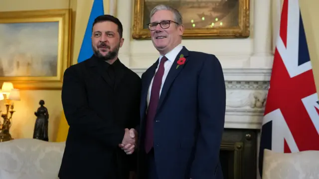 Volodymyr Zelensky (L) shakes hands with Keir Starmer (R) insider Number 10, a Ukrainian flag and a Union flag behind them to the sides of a marble fireplace