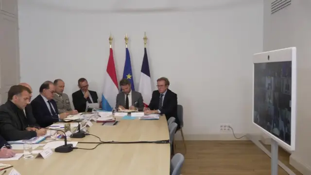 A conference table where Emmanuel Macron is sat, it faces a monitor with the screens of other world leaders who are on a video call