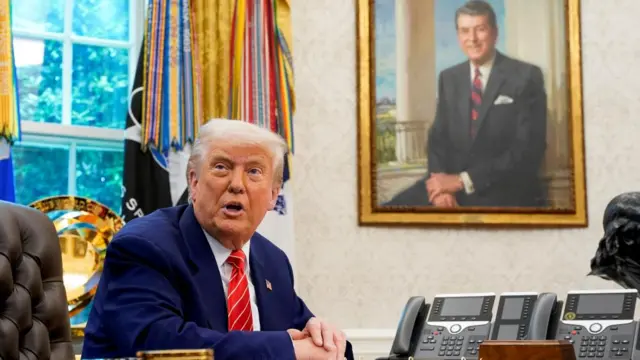 Trump seen sitting at his desk in the Oval Office with a portrait of President Reagan behind him