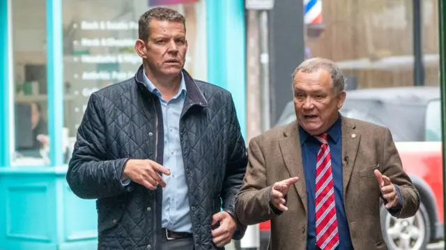 Rhun and Lindsay walking side by side on a streets. they are deep in conversation, with whittle gesticulating with his hands.