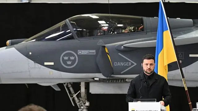 A man in a dark suit stands in front of a Ukrainian blue and yellow flag and an air plane