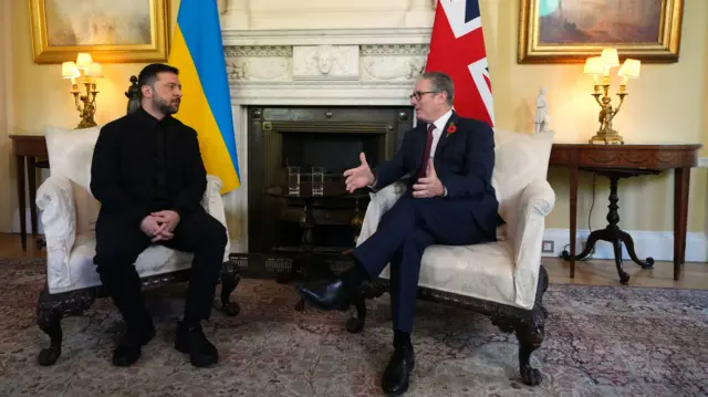 Volodymyr Zelensky (L) and Keir Starmer (R) sit down on white brocade chairs inside Number 10, a marble fireplace between them