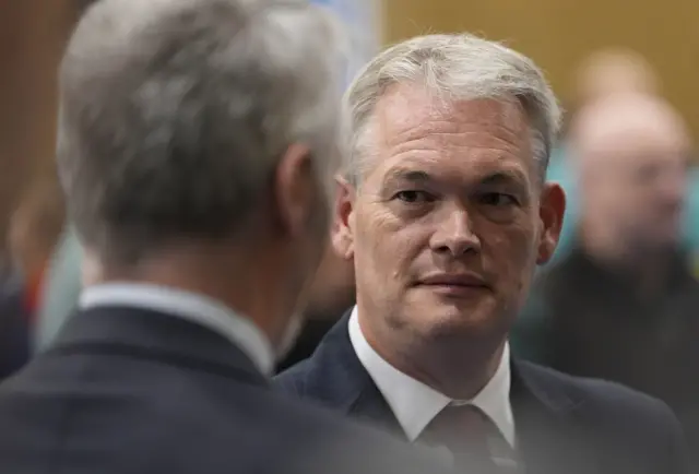 Tunnicliffe speaks to another man in the count. the back of the person he's talking to is visible. Tunnicliffe is wearing a dark suit and red tie