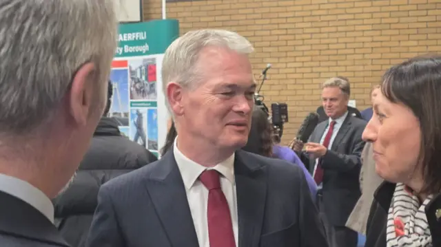 Richard Tunnicliffee wearing a black suit, red tie and white shirt between two people in a counting hall.