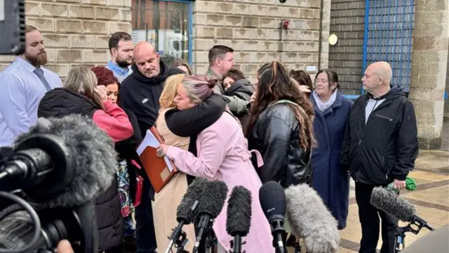 A group of people gathered and are hugging outside a court building in front of lots of microphones on stands