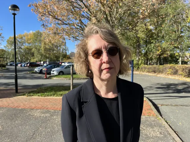 Miranda stands in a car park with her arms behind her back, with a sombre expression. She has short wavy hair, and wears a black blazer, black top and dark sunglasses.