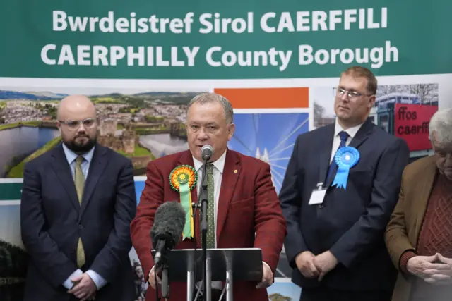 Whittle speaks at the podium giving his acceptance speech. He is flanked by the Reform candidate Powell and the conservative candidate Potter