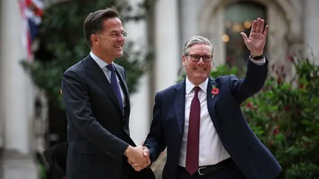 Mark Rutte shakes the hand of Keir Starmer who raises up his other hand in a wave