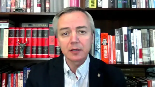 A man in a black jacket and white shirt sits in front of a bookshelf