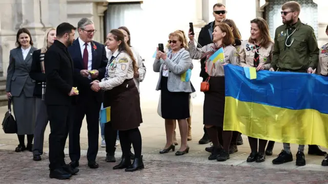 Zelensky shakes hands with a supporter, with Starmer standing nearby. A group of supporters holding a large Ukrainian flag is standing near them.
