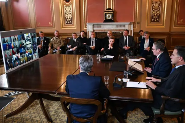 Four men and one woman sit at one end of brown table, at the other end is a TV screen with around 25 people on it as part of a video call. A handful of people in formal clothing sit and watch.