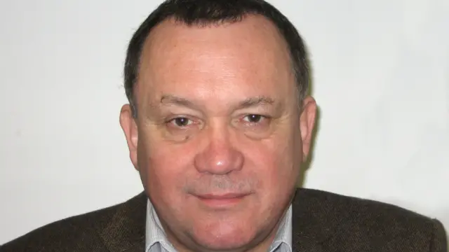 A head shot of Lindsay looking at the camera. He is younger and his hair is darker. he is wearing a dark brown jacket.