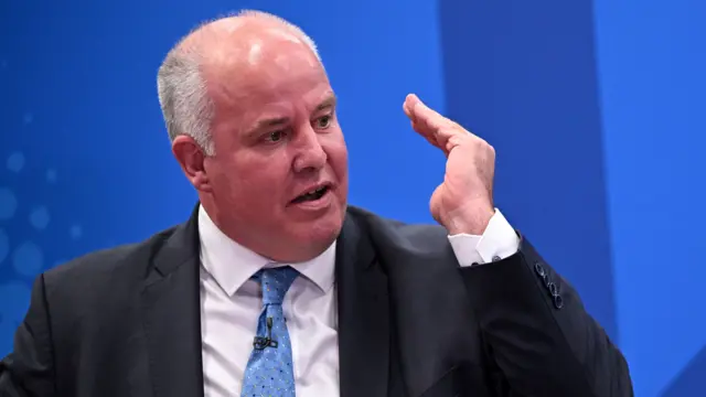 Photograph of Andrew RT Davies, wearing a black suit, blue tie and white shirt. He has silver hair on the sides of his head and holds his hand up to his face. In the background is a blue screen.