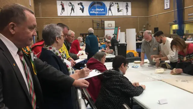 Lindsay stands in profile looking at a bank of desks where people are counting ballots. His hand is resting on a chair and he looks concerned.